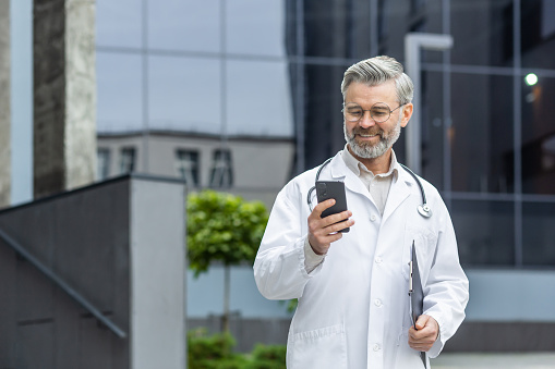 A male senior doctor in a white coat, with a stethoscope and a pack in his hands stands outside the clinic and uses the phone, writes a message, talks on a video call, smiles.
