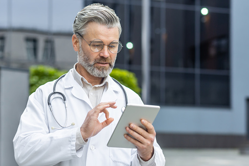 Online medicine. A senior male doctor stands outside the clinic in a white coat and holds a tablet in his hands. Chats with patients, consults on a video call, points to the camera with his fingers.