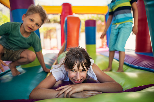 Kids having extreme fun in inflatable bouncy castle playground. Sunny summer day. The girl is lying on the front and smiling at the camera while the boys are jumping.\nNikon D810