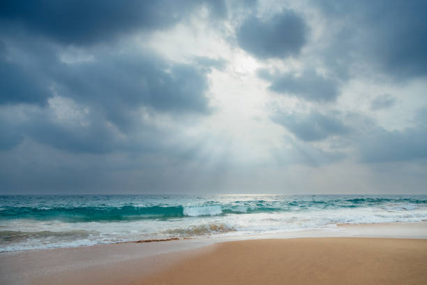 A gorgeous low-angle view of a sandy ocean beach with turquoise waves under a dramatic cloudy sky with sunlight through it.