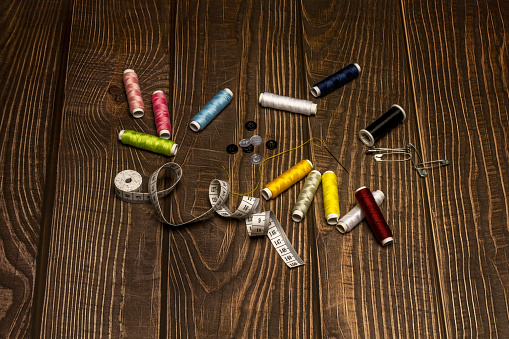 A composition of sewing items with various spools of colored thread, a group of safety pins, buttons, a tape measure, scissors, a needle with threaded thread on a dark wooden table