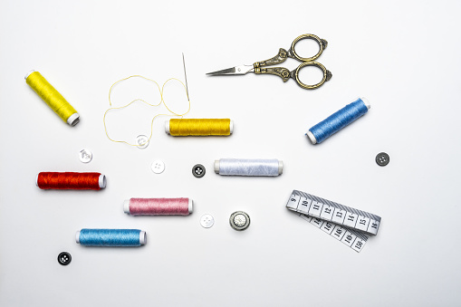 View of black and white buttons, needle with threaded yellow thread, various colors on spools of thread on white background and vintage sewing scissors