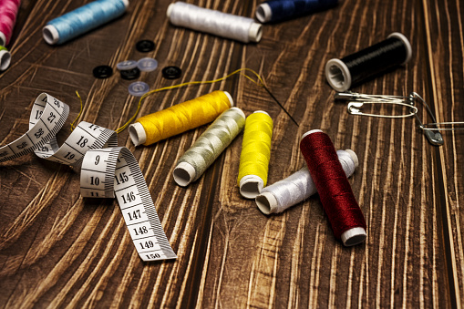 A composition of sewing items with various spools of colored thread, buttons, tape measure, scissors, a needle with threaded thread on a wooden table