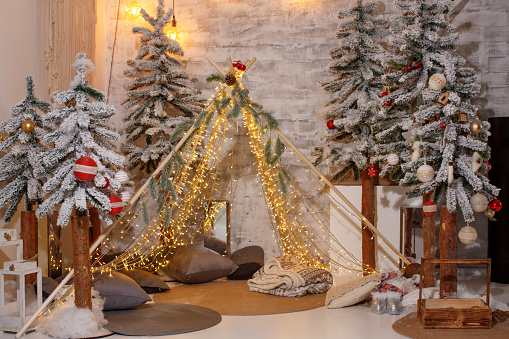 Tepee of luminous garlands against the backdrop of a brick wall and decorated Christmas trees
