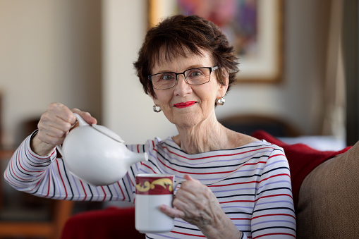 Senior woman sitting at home pouring tea from a teapot.