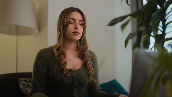 A young gen z woman is sitting on a sofa burning incense and watching online videos for meditation.