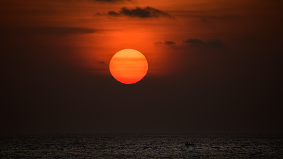 Big ball of the orange sun setting behind the ocean horizon, beautiful sunset colors in the sky. Solitary silhouette fishing boat sails in the ocean.