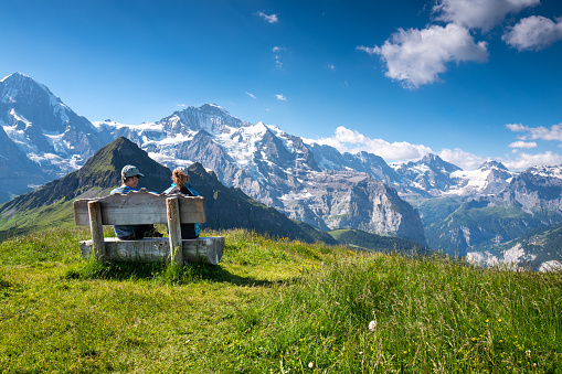 Mannlichen, Switzerland - July 3, 2022: Young couple enjoying the view of Jungfrau and Eiger in the Swiss Alps from a bench on the mountain Mannlichen on a warm summer day