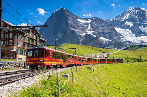 Kleine Scheidegg, Bernese Oberland, Switzerland - July 3, 2022:  The famous Red Jungfraubahn train that travels to and from Jungfraujoch and Kleine Scheidegg, passing through the Mountain Eiger and making it the highest railway line in Europe. Eiger and Jungfrau mountain peaks in the background.