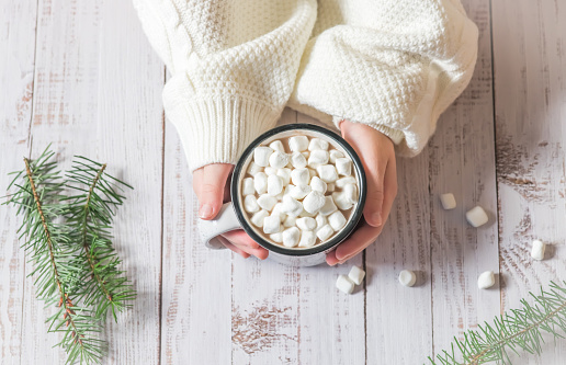 Girl hands in white knitted sweater holding a cup of hot chocolate with marshmallows on top  during winter holidays on a wooden background.