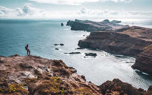 Description: Backpacker woman enjoys panoramic view from a steep cliff overlooking the sea and the rugged foothills of Madeira's coast in the morning. Ponta do Bode, Madeira Island, Portugal, Europe.