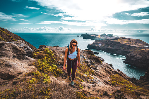 Description: Backpacker woman enjoys hike along a steep cliff overlooking the sea and the rugged foothills of Madeira's coast in the morning. Ponta do Bode, Madeira Island, Portugal, Europe.