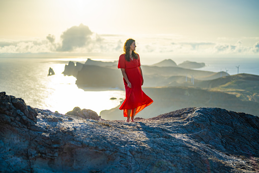 Description: Woman in red dress enjoys panoramic view from steep cliff over seascape and along rugged foothills of Madeira coast at sunrise. Ponta do Bode, Madeira Island, Portugal, Europe.