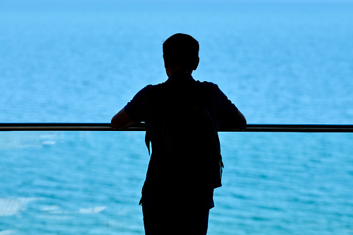 Silhouette of young man leaned on transparent glass handrail on above blue sparkling sea background, beautiful touristic point of view, rear view. Man silhouette contemplating fresh air seascape