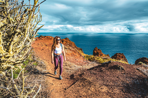 Description: Backpacker woman enjoys hike along a steep cliff overlooking the sea and the rugged foothills of Madeira's coast in the morning. Ponta do Bode, Madeira Island, Portugal, Europe.