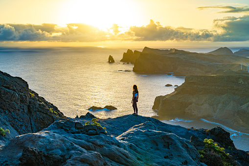 Description: Woman enjoys panoramic view from view point on a steep cliff over the seascape and along the rugged foothills of Madeira coast at sunrise. Ponta do Bode, Madeira Island, Portugal, Europe.