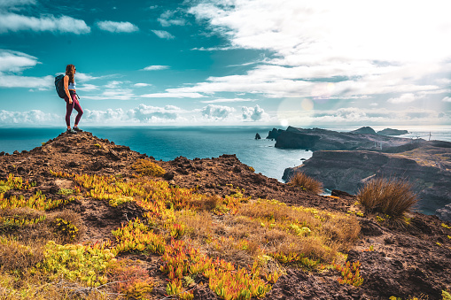 Description: Backpacker woman enjoys panoramic view from a steep cliff overlooking the sea and the rugged foothills of Madeira's coast in the morning. Ponta do Bode, Madeira Island, Portugal, Europe.