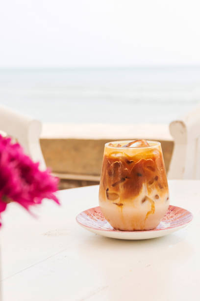 glass of iced caramel macchiato on white wooden table with blue sea on background. - caramel latté coffee cafe macchiato imagens e fotografias de stock