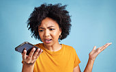 Shot of a young woman standing against a blue background in the studio and looking confused while using her cellphone