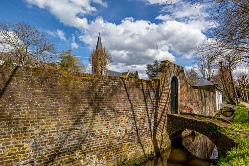 Old brick wall with a gate and small bridge along canal, bell tower of St. Michael's Abbey Church in background