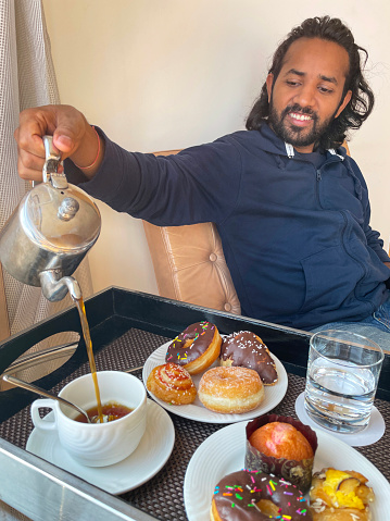 Stock photo showing close-up, elevated view of a wooden tray containing a white cup and saucer with teaspoon, a metal pot of freshly brewed coffee, plates of doughnuts and pastries and drinking glass of water as part of room service breakfast.