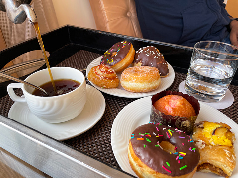 Stock photo showing close-up, elevated view of a wooden tray containing a white cup and saucer with teaspoon, a metal pot of freshly brewed coffee, plates of doughnuts and pastries and drinking glass of water as part of room service breakfast.