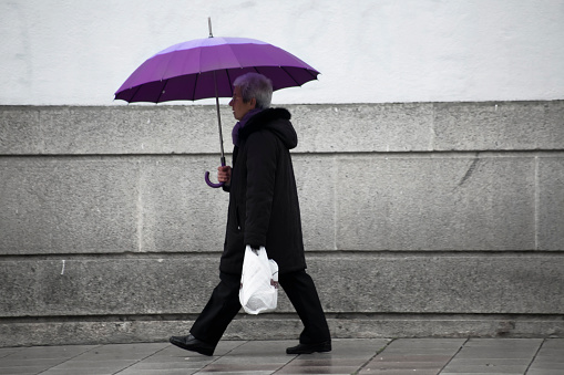 Lugo, Spain - April 11, 2012 Side view of senior woman walking on a street sidewalk carrying shopping plastic bag and umbrella in a rainy day. Lugo city, Galicia, Spain.