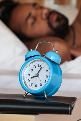 Stock photo showing close-up view of a blue metal case retro alarm clock with double bell on night stand. An Indian man can be seen lying in the double bed, next to the beside table, sleeping.