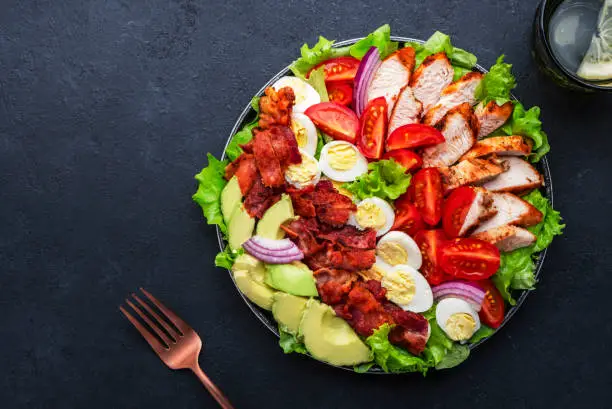 Cobb salad with chicken fillet, tomatoes, eggs, bacon, avocado and lettuce, dark table background, top view. American cuisine dish