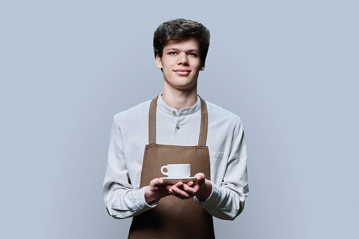 Young smiling male barista waiter employee in apron posing on gray studio background with cup of coffee. Coffee house shop, restaurant, cafe worker looking at camera