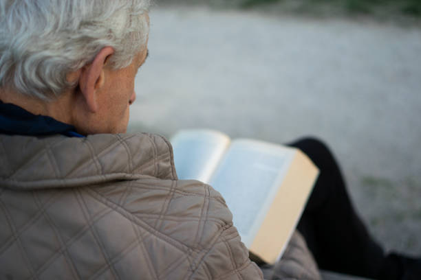 Old man with gray hair reading a book sited on a bench outside in park