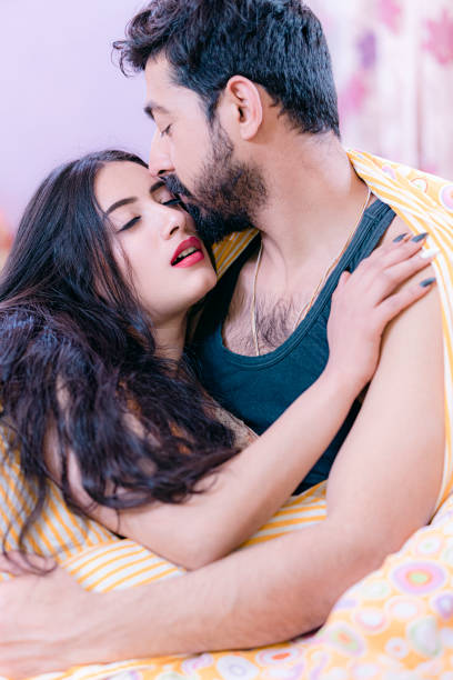 Handsome man embraces his beauiful girlfriend. An Asian/Indian mid-adult man wearing a sleeveless vest embraces his beautiful young adult girlfriend in the bedroom. They sit together, wrapped in the same quilt. indian couple kiss stock pictures, royalty-free photos & images