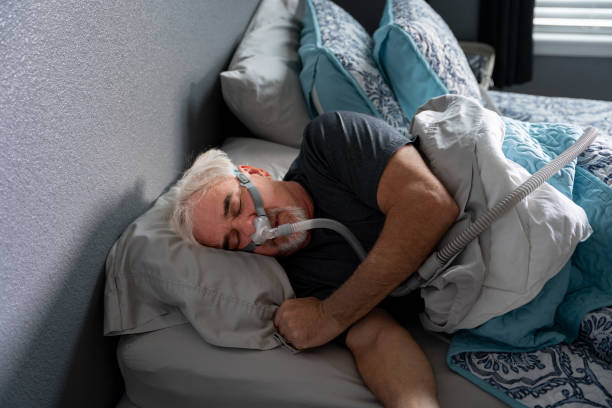 A 60-year-old man sleeps with a CPAP machine in bed to treat his sleep apnea, a condition where breathing repeatedly stops and starts during sleep. stock photo