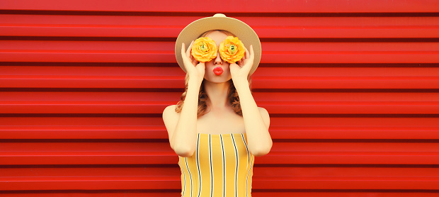 Summer portrait of happy young woman covering her eyes with flower buds as binoculars looking for something wearing round straw hat on red background