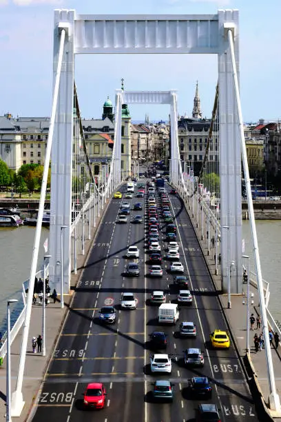 the Elizabeth bridge in Budapest. perspective view. white steel frame with heavy suspension cables. car traffic on the highway. old European architecture. boats on the Danube river. travel and tourism