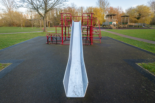 Tall, high, skinny, stainless steel silver slide coming off metal play structure in children's playground with grass, trees and soft safe floor covering. Image taken from bottom of slide looking up.