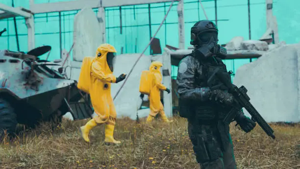 Workers in radiation protection suits are escorted through a dangerous area by soldiers holding weapons. Maybe they are worried about a virus, maybe it is radiation or some sort of biochemical warfare. A destroyed building is in the background.