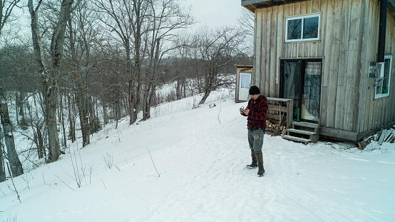An aerial shot of a man flying his drone in the snow with a log cabin behind him