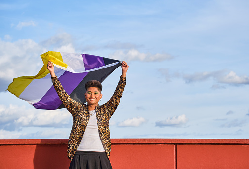 asian gender non binary person smiling holding gender non binary representative flag blowing in the wind with blue sky background