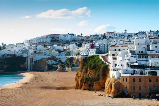 View over Albufeira in the Algarve Province of Portugal on a sunny day. Whitewashed buildings contrasting with the blue sky and a few light clouds. Large expanse of sand of the Praia de Pescador (Fisherman's Beach) in the foreground. This town on the south coast of Portugal is a popular tourist destination all the year round on account of its mild winters, warm and sunny summers and beautiful beaches.