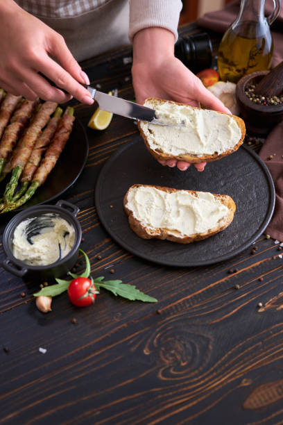istock Making breakfast toasts - woman smearing cream cheese on a grilled bread over kitchen table with Asparagus wrapped with bacon and spices on a plate 1484244870