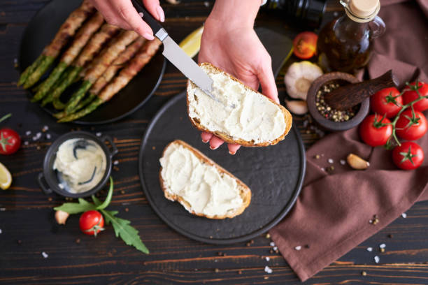 istock Making breakfast toasts - woman smearing cream cheese on a grilled bread over kitchen table with Asparagus wrapped with bacon and spices on a plate 1484244888