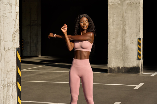 Young adult confident beautiful fit sporty slim active black African ethnic woman runner model wearing sportswear doing warming up exercise training standing in urban location outdoors.