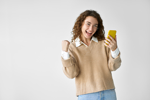 Young happy excited pretty woman student feeling winner holding cellphone using mobile phone winning game app prize, receiving great job offer celebrating standing isolated at white background.