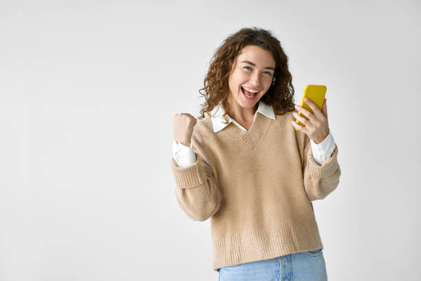молодая счастливая женщина чувствует себя победителем, используя мобильный телефон, стоящий на белом фоне. - scholarship holder стоковые фото и изображения