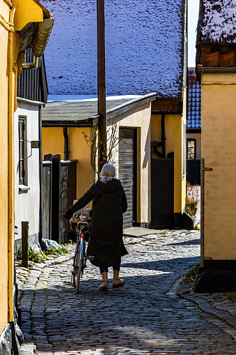 Dragor, Denmark March 31, 2023 A senior woman walking with a bicycle on a cobblestoned street in the sprintime with snow on the roofs.