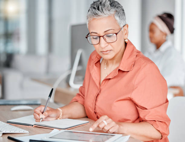 Tablet, book and business woman writing, research or internet browsing in office workplace. Technology, notebook and senior female employee with touchscreen for taking notes and planning strategy.