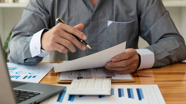 istock businessman in shirt holding documents Man working with documents, financial graphs and bills sitting at desk using laptop at work analyzing accounting finances remotely. 1484260325