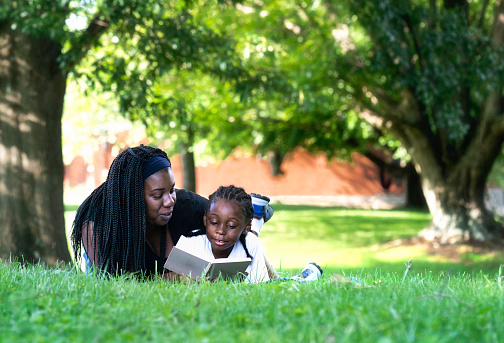 Mom teaches her 6 year old son to read a book outdoors under a tree on a sunny day, Indiana, USA