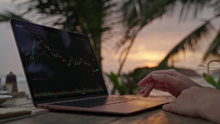 Female cryptocurrency trader at laptop checking candlestick charts online working remotely at outdoor tropical seaside cafe. Woman crypto broker analyses stock exchange rates and bets by ocean closeup